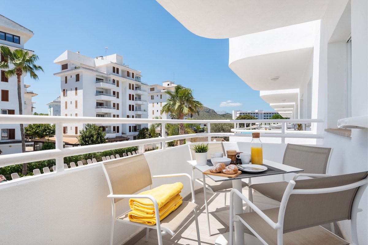 A private terrace is presented with outdoor seating, featuring a table set for dining. Bright yellow towels are neatly arranged on a chair, while a refreshing drink and light breakfast are displayed. The view includes nearby buildings and palm trees under a clear blue sky.