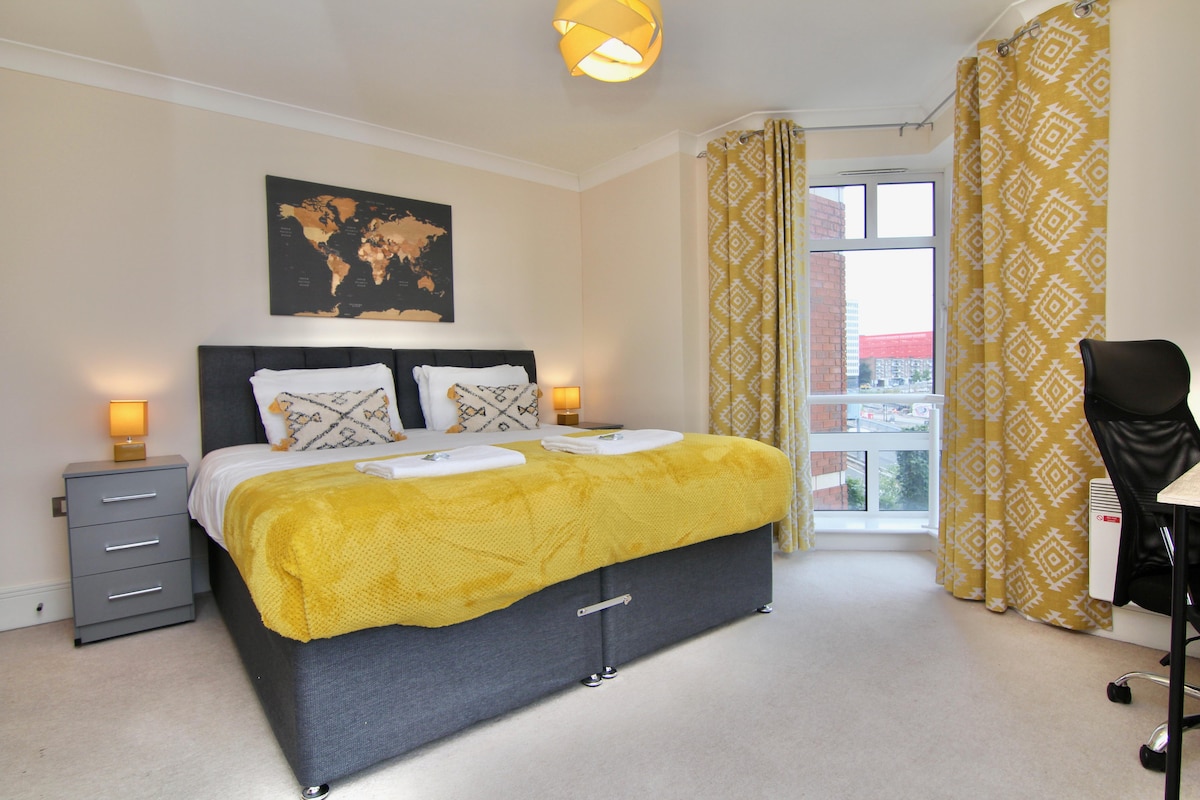 A spacious sleeping area features a large bed with a soft yellow spread. Two bedside lamps illuminate the space, complemented by patterned curtains. A world map art piece adorns the wall, and a work desk with a chair is positioned nearby, allowing for a functional workspace.