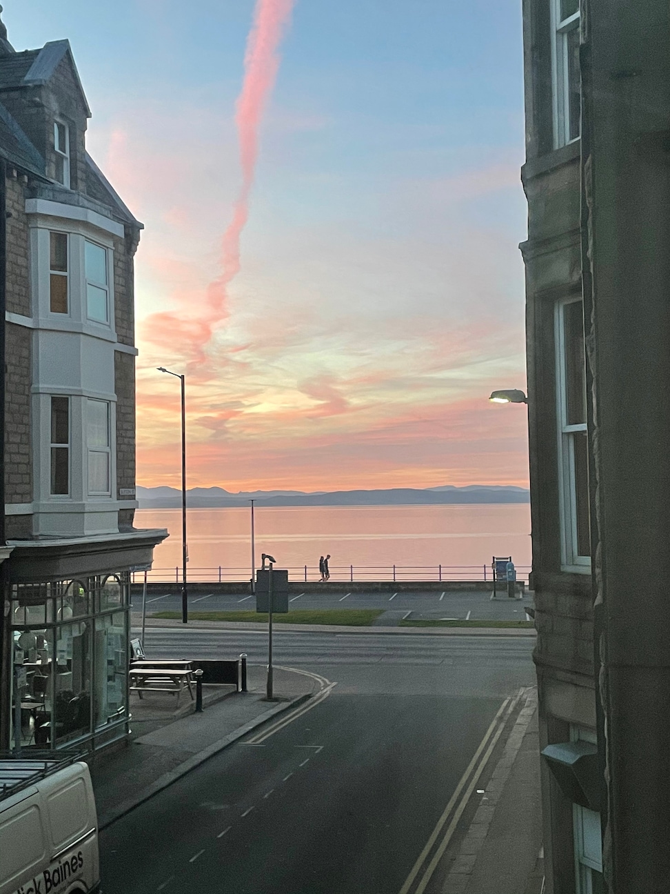 A stunning view of the sea at sunset is captured, displaying soft pastel colors in the sky. The waterfront is visible, along with people walking along the promenade, framed by nearby buildings and streetlights.
