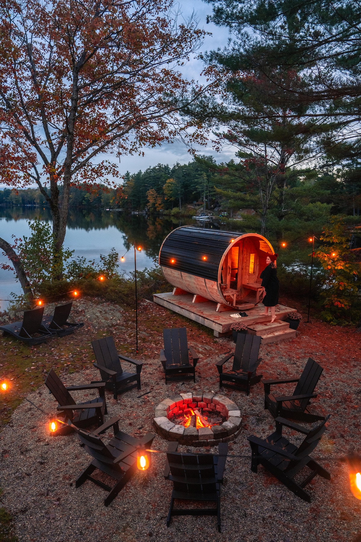 A circular cedar barrel sauna is nestled by the lake, surrounded by a circular seating area of black adirondack chairs. A fire pit with glowing embers sits at the center, while nearby torches provide soft lighting against a backdrop of autumn foliage.