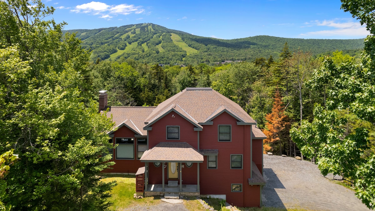 The exterior of the home is showcased amidst lush greenery, with a rustic red façade and a welcoming entrance. Mount Snow is visible in the background, accentuating the scenic landscape. The surrounding trees create a serene and private atmosphere.