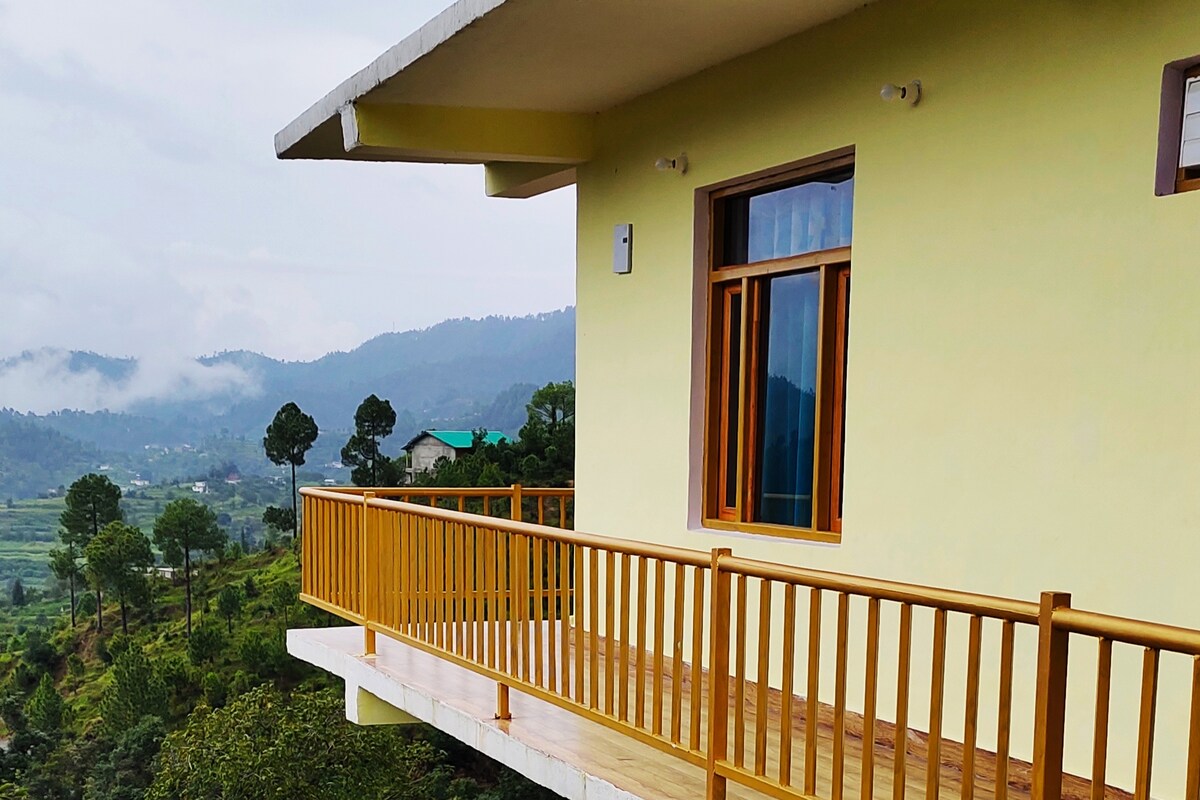 A wooden balcony extends from the guest room, providing a panoramic view of the surrounding hills and valleys. Large windows allow natural light to fill the space, while the simplistic architecture blends harmoniously with the natural landscape.