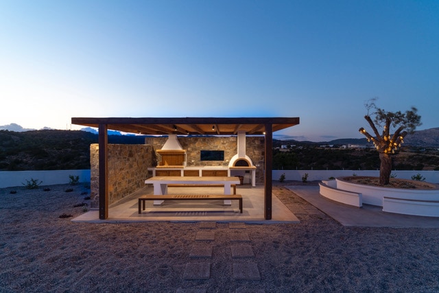 An outdoor barbecue area is highlighted, featuring a stone structure with a wooden roof. A large dining table and benches are positioned underneath, accompanied by an outdoor oven. Surrounding gardens are visible, providing an inviting setting for cooking and dining under the evening sky.