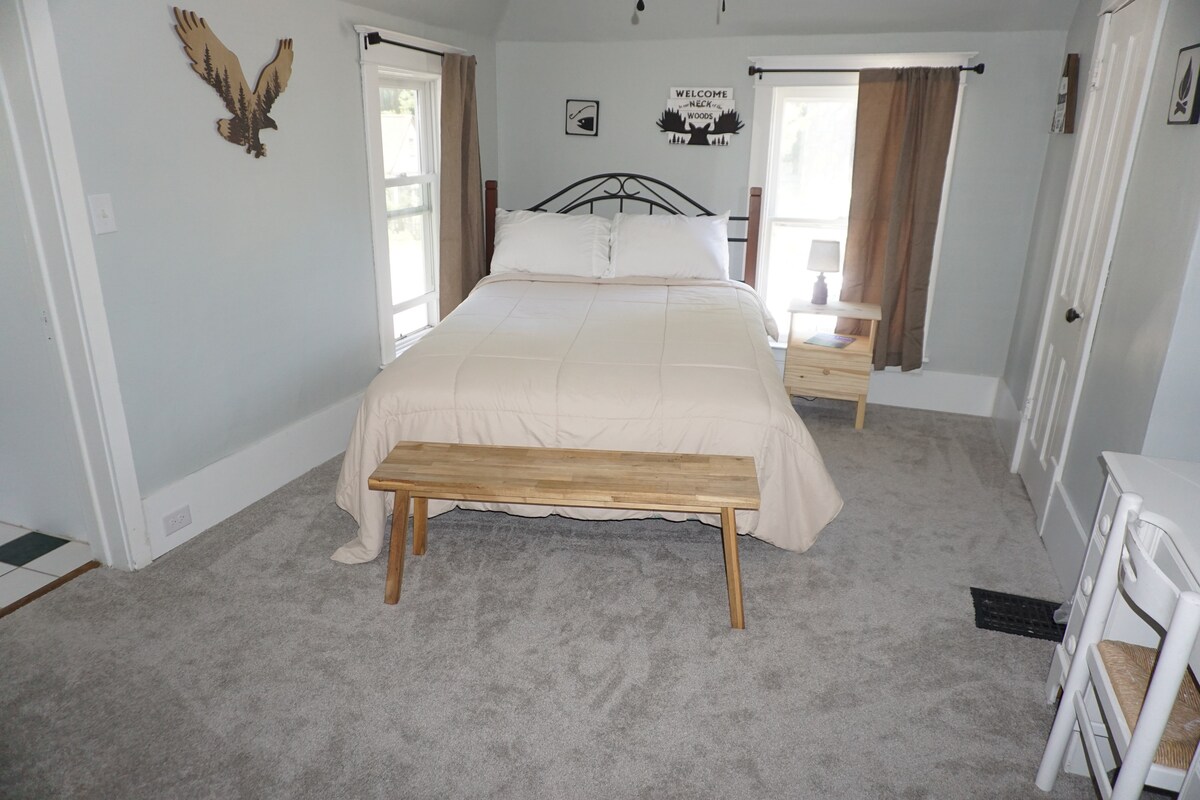 The room features a queen size bed with a light-colored comforter, positioned against a wall with decorative wall art. A wooden bench sits at the foot of the bed, and a bedside table with a lamp is present. Windows with light curtains allow natural light into the space.