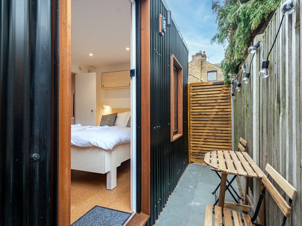 A private outdoor space is visible, featuring a wooden table and stools, positioned against a fence. The entrance to the cabin reveals a well-lit sleeping area with a double bed. Natural light highlights the clean lines and contemporary design of the cabin.