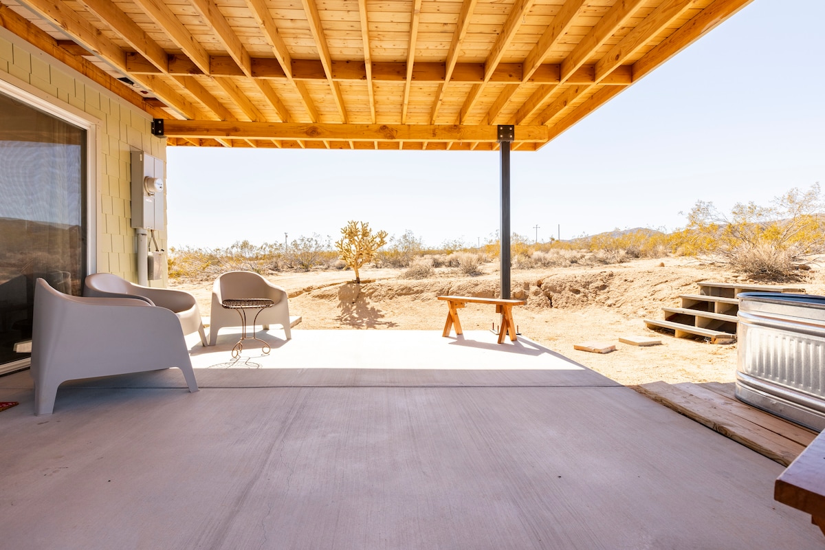 A covered outdoor patio area features minimalist seating arrangements with two chairs and a wooden bench. The desert landscape is visible beyond, showcasing dry terrain and sparse vegetation under a clear blue sky.