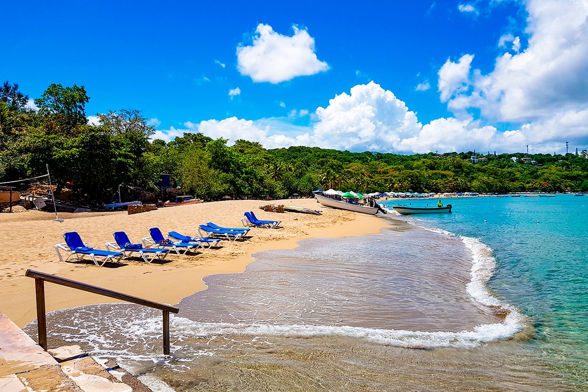 A sandy beach is bordered by clear blue water, with several lounge chairs positioned under the sun. Lush greenery lines the background, while boats can be seen floating nearby. The sky features a mix of clouds and sunlight, contributing to a serene coastal atmosphere.