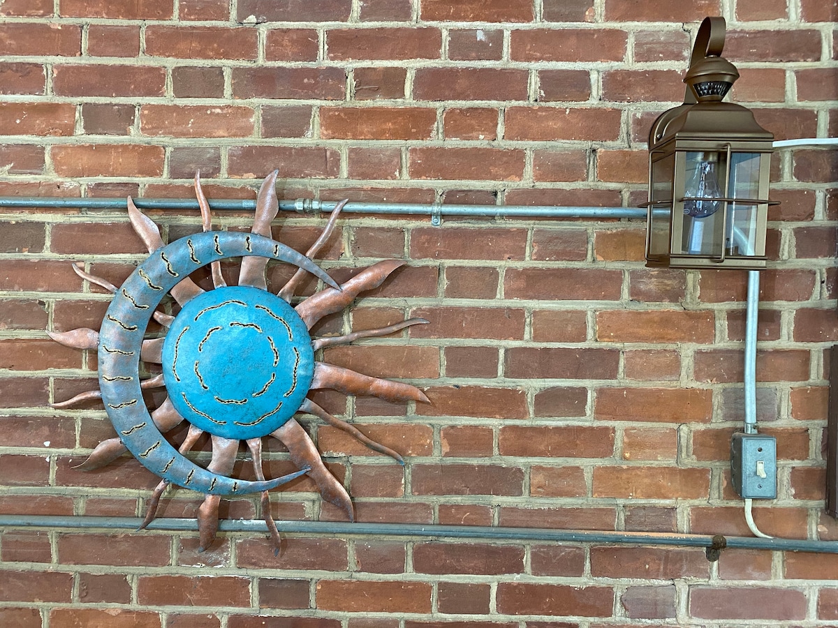 A decorative sun sculpture with a bright blue center is mounted on a brick wall, surrounded by rays in earthy tones. A vintage-style lantern hangs nearby, adding an element of warmth. The exposed brick and utility pipes contribute to the unique character of the space.