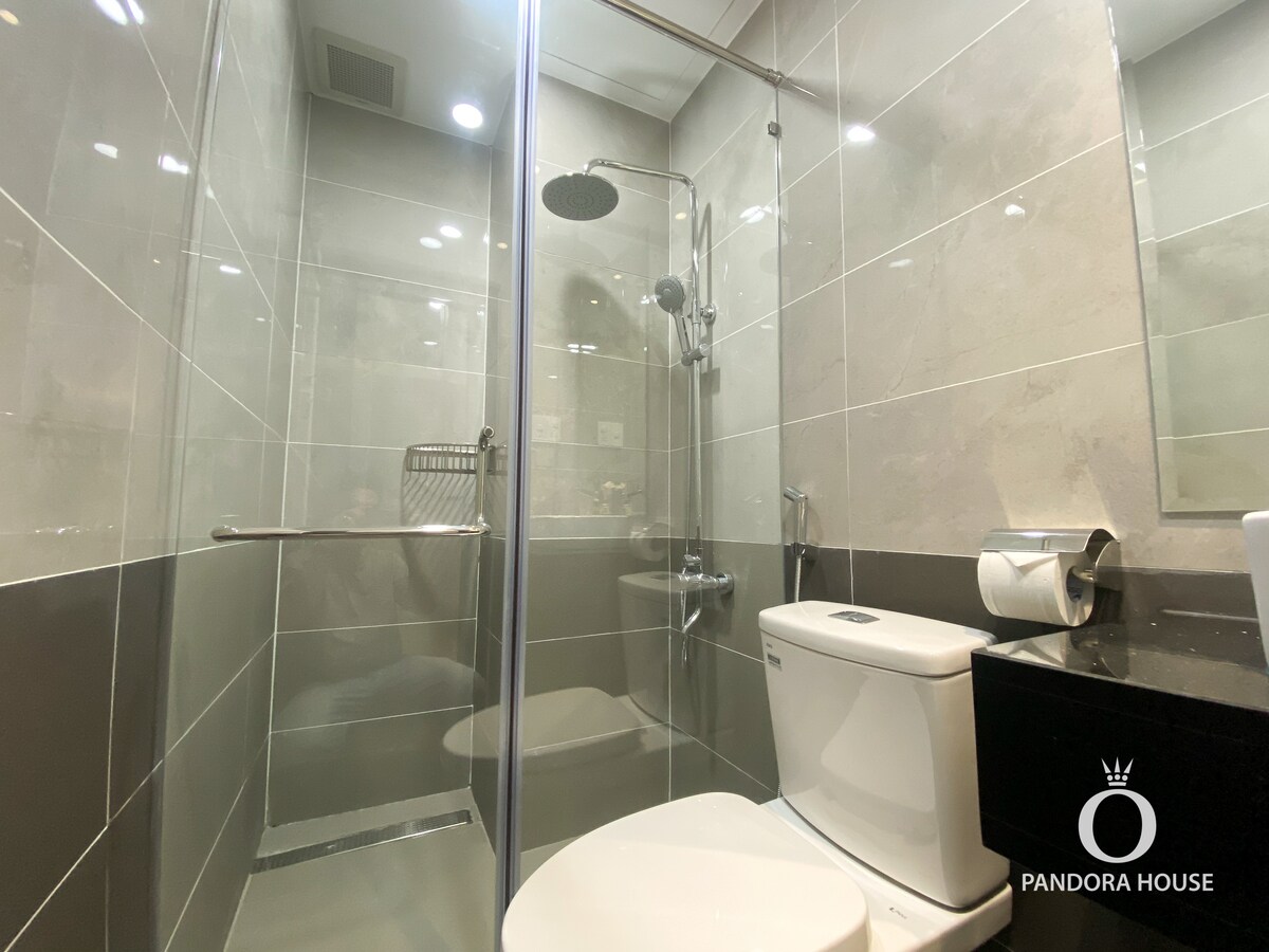 A modern bathroom features a glass shower enclosure with a rainfall showerhead and chrome fixtures. A sleek white toilet is positioned beside a black countertop with essential toiletries. Gray tiles create a clean and polished look throughout the space.