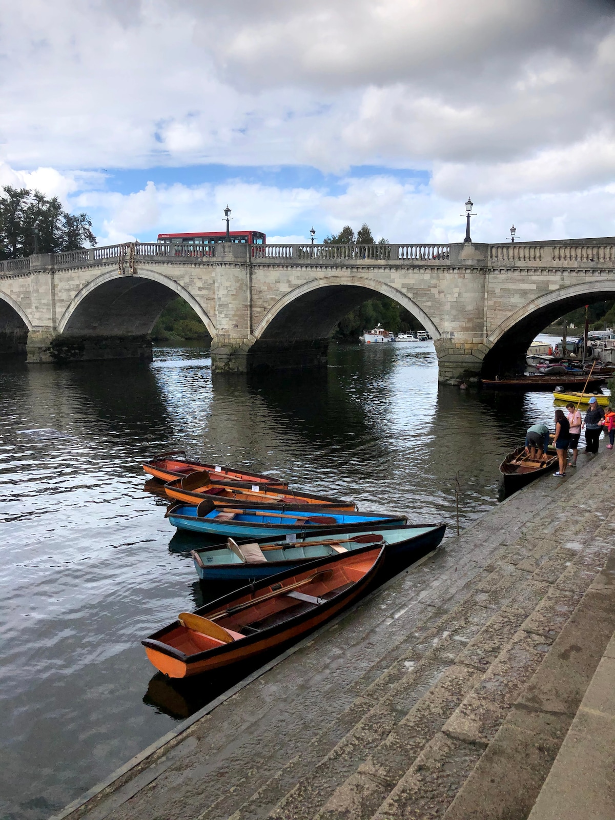 A serene riverside scene features several wooden boats floating gently on the water, displaying a mix of colors including blue and orange. A stone bridge arches over the river, while a pathway lined with trees invites leisurely strolls along the bank.