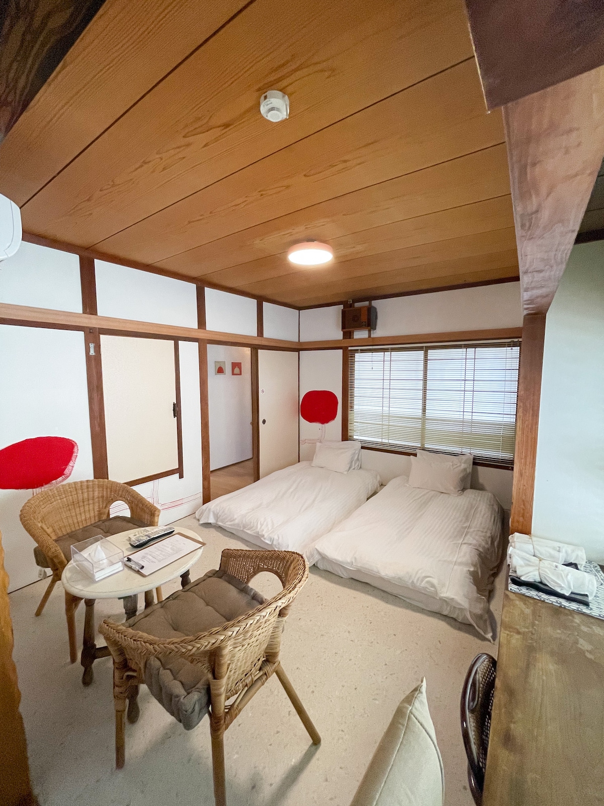 The guest room features two single mattresses laid on a simple floor. A table with chairs is positioned nearby, complementing the spacious layout. Natural light enters through a window adorned with traditional blinds, while wooden beams create a warm ambiance.
