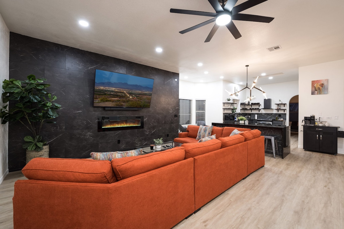A spacious living area features a large orange sectional sofa facing a mounted flat-screen TV. A decorative plant is positioned to one side, while an elegant fireplace is integrated into the wall. The open layout extends towards the kitchen and dining area, enhancing the overall ambiance.