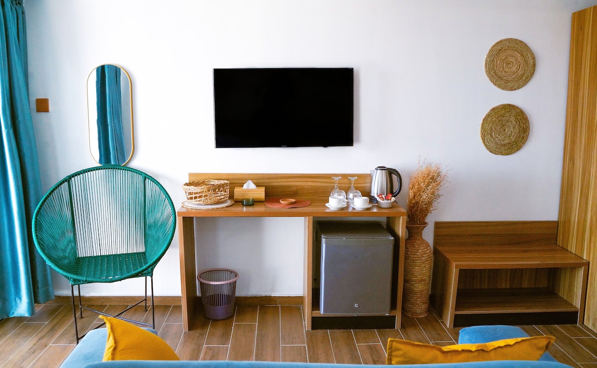 A modern seating area is highlighted by a green chair and a wooden console featuring a kettle and refreshments. A flat-screen television is mounted on the wall, complemented by woven wall decor and natural elements, creating a simple yet stylish environment.