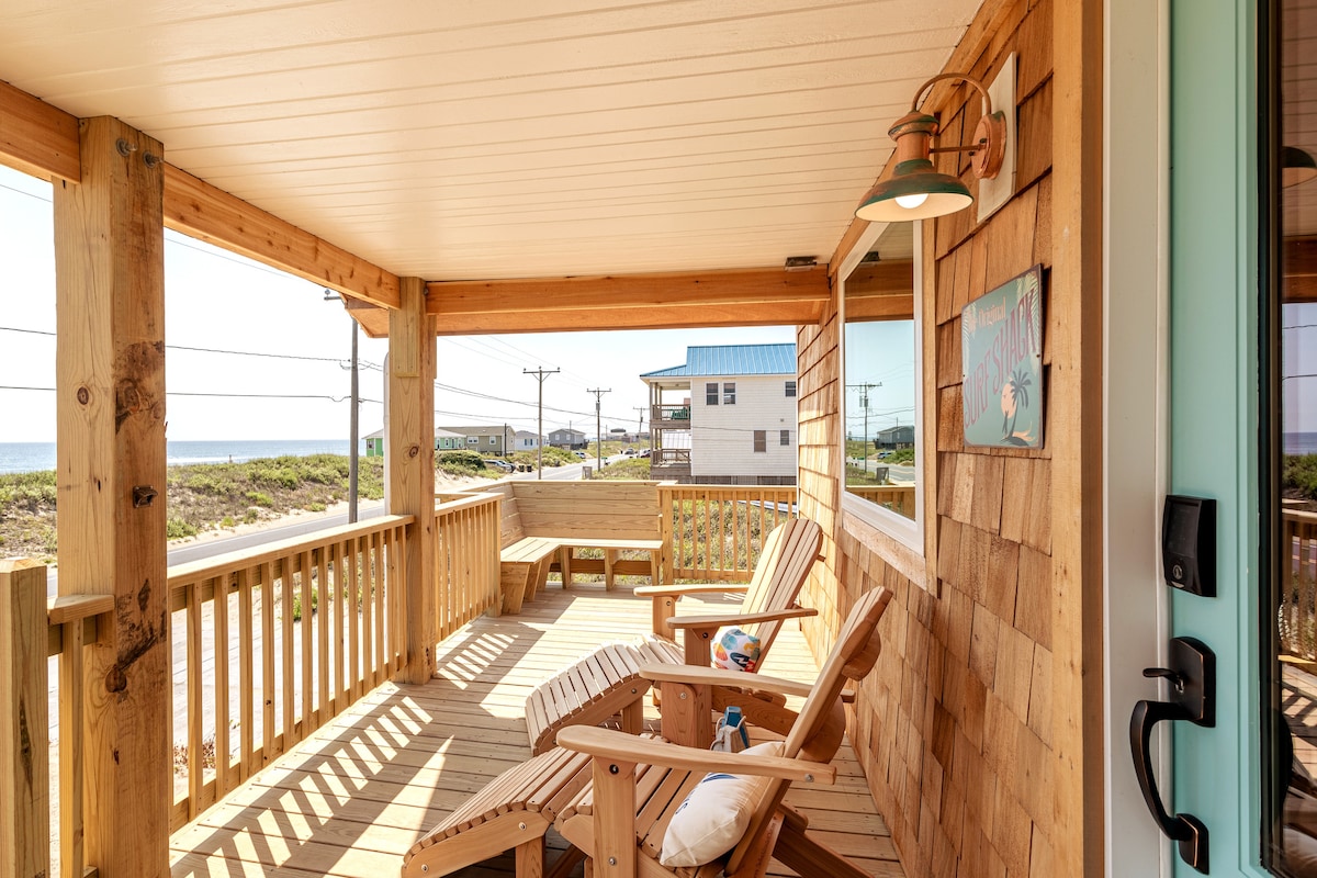 A spacious covered porch features two wooden lounge chairs positioned for ocean views. Natural wood siding and a light fixture complement the inviting outdoor space. The beach is visible in the distance, enhancing the relaxed coastal atmosphere.