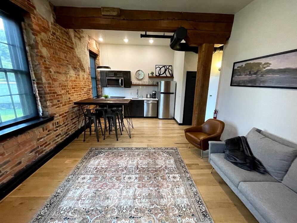 An open-concept living area showcases exposed brick walls and hardwood floors beneath wooden beams. A modern kitchen with a dining table and four chairs is visible. A light-colored sofa rests on a patterned rug, and a framed landscape hangs on the wall near a large window.