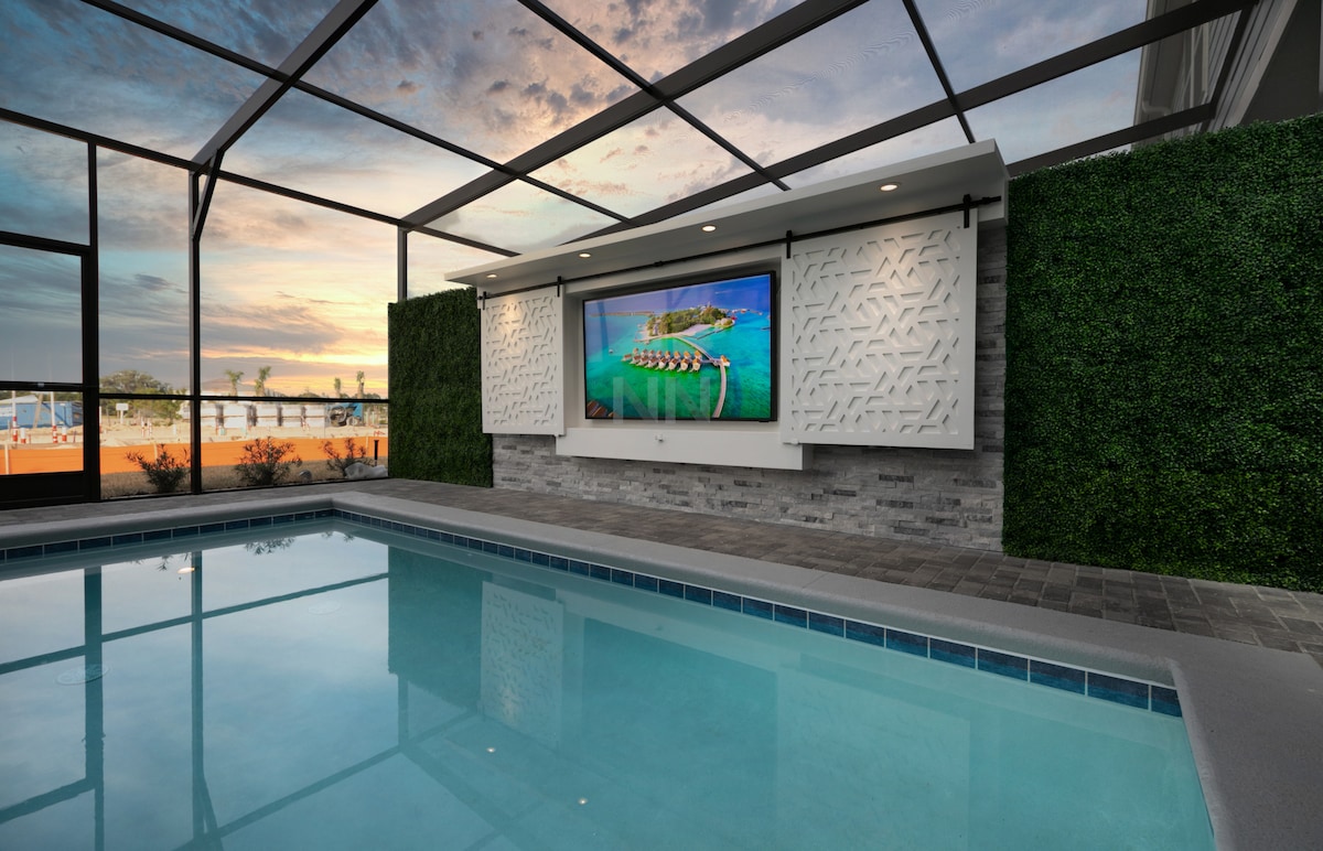 An inviting outdoor pool area is enclosed with a screened lanai, featuring a mounted large TV above a stone wall. Lush greenery provides a backdrop, while the pool water reflects the soft evening light.