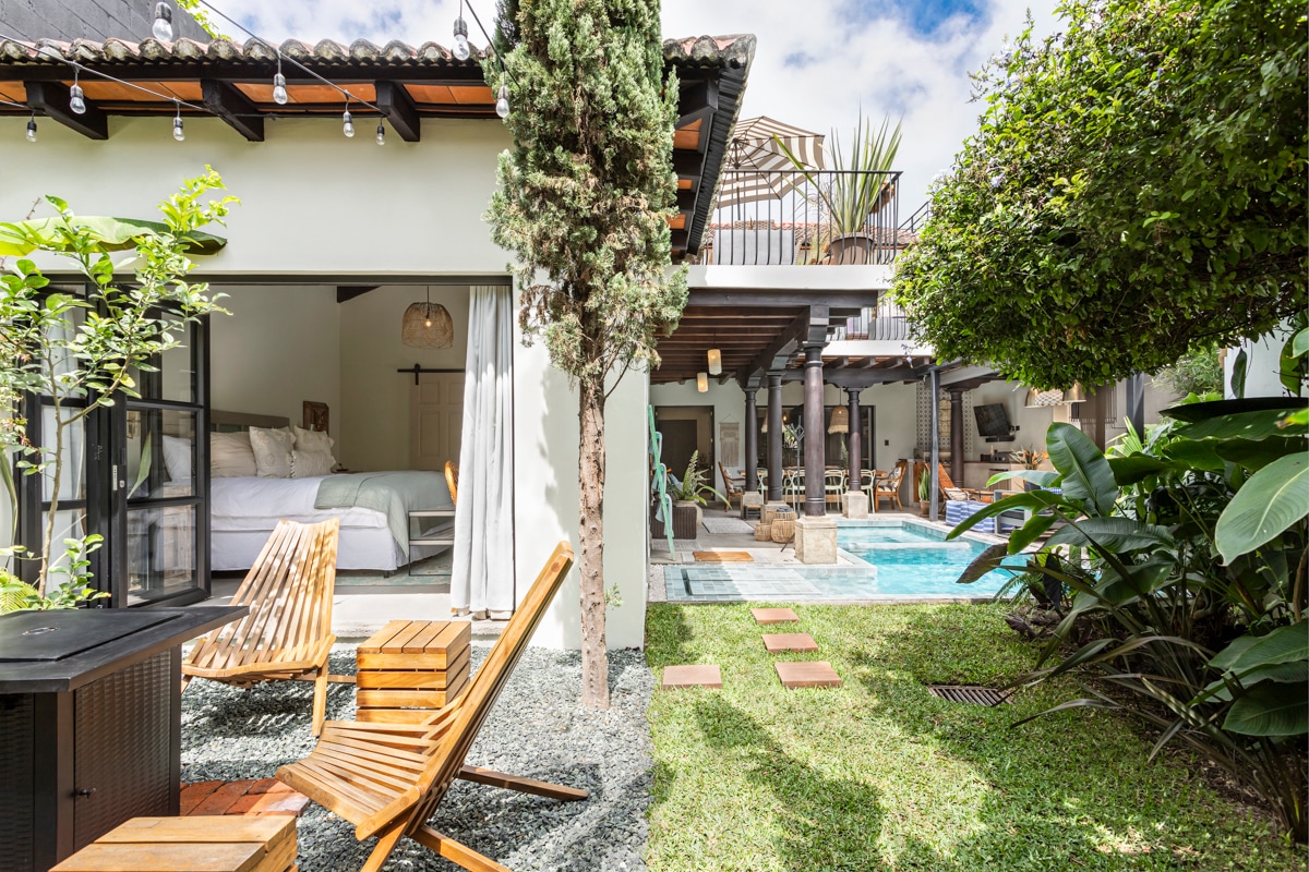 A landscaped garden features lush greenery surrounding a sparkling swimming pool. A wooden deck with two lounge chairs is positioned nearby, offering a relaxing space. The open-concept area reveals a light-filled interior, blending seamlessly with outdoor elements.