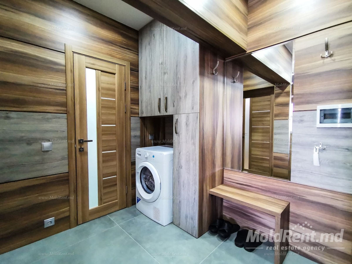 A functional laundry area is featured, with a washing machine positioned against wood-paneled walls. A small bench is located nearby, complemented by hooks for hanging coats. Full-length mirrors add depth, while a wooden door leads to the rest of the apartment.