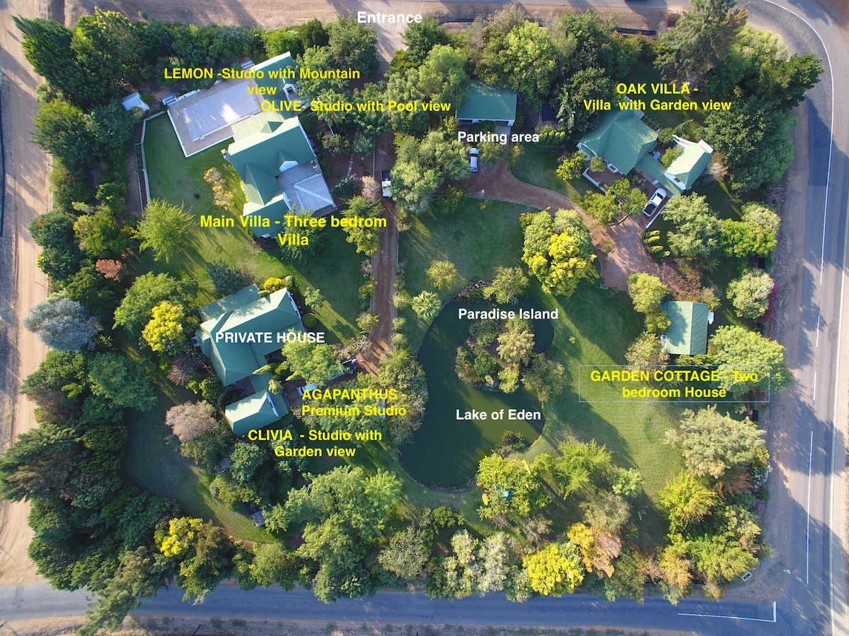 An aerial view of the property shows a layout of various accommodations including the Lemon Studio, Oak Villa, and Garden Cottage. The lush garden surrounds a tranquil lake, with clearly marked areas for parking and the entrance. Pathways connect the units, creating an inviting atmosphere.