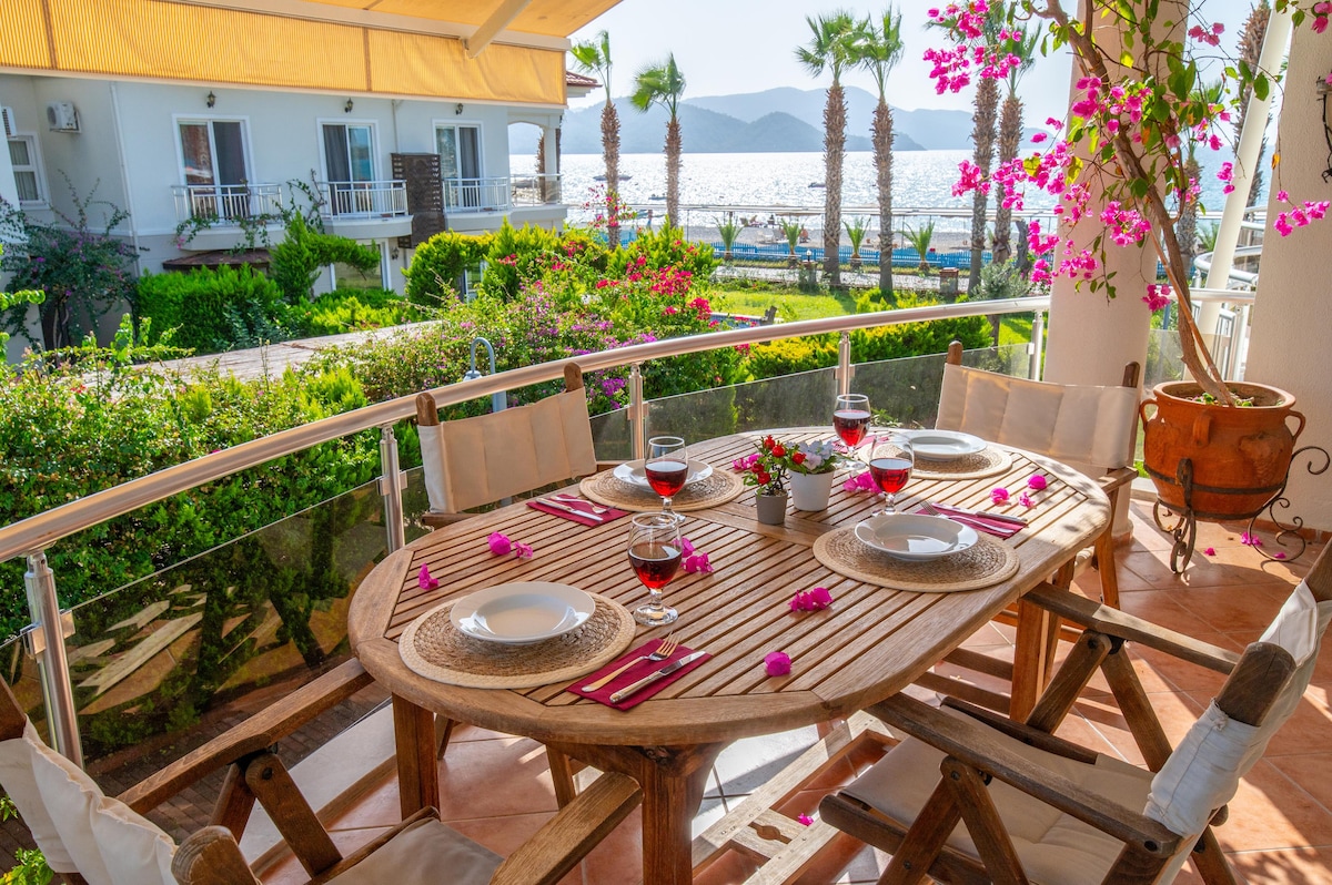 An outdoor dining area features a wooden table set for a meal, adorned with wine glasses and colorful utensils. The space is surrounded by lush greenery and flowering plants, offering a sea view in the background. A bright atmosphere is created by the sunlight filtering through an awning.