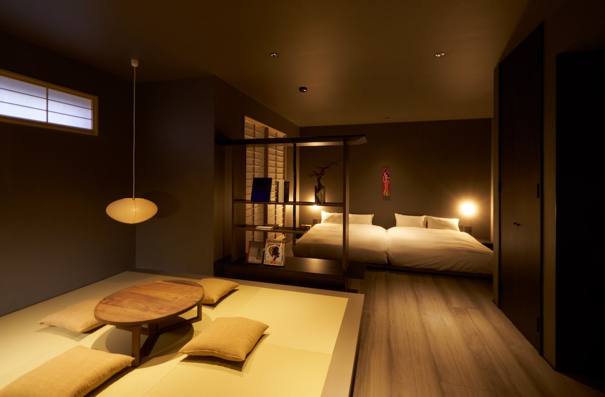 A spacious room features a tatami seating area with a low wooden table and cushions. Beyond, two beds are arranged in a minimalist style. Soft lighting creates a calming ambiance, complemented by subtle decorative elements on the walls.