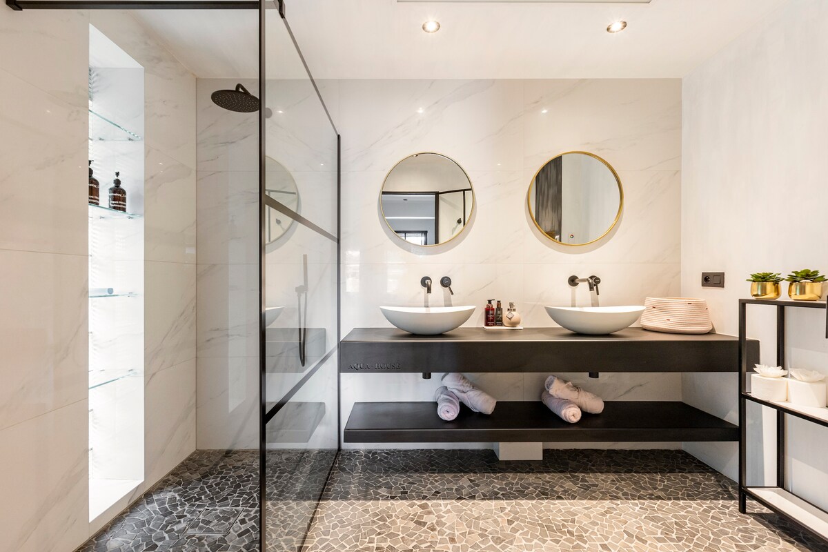 A modern bathroom features a spacious shower enclosure with a rain shower head. Dual circular mirrors are mounted above a sleek wooden vanity with two washbasins. Textured flooring complements the minimalist design, while neatly rolled towels are placed on the vanity.