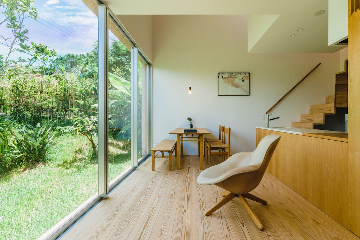 A spacious dining area features a wooden table and chairs, complemented by a lounge chair. Large glass doors provide a view of the lush greenery outside. Natural light enhances the warm wooden flooring and the simplicity of the interior design.