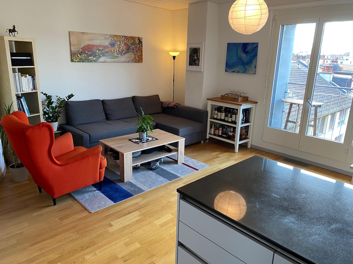 The living room features a grey sectional sofa, an orange armchair, and a coffee table set atop a multi-coloured rug. A floor lamp stands beside the sofa. Light enters through large windows that provide views of the outside, with a small balcony visible.