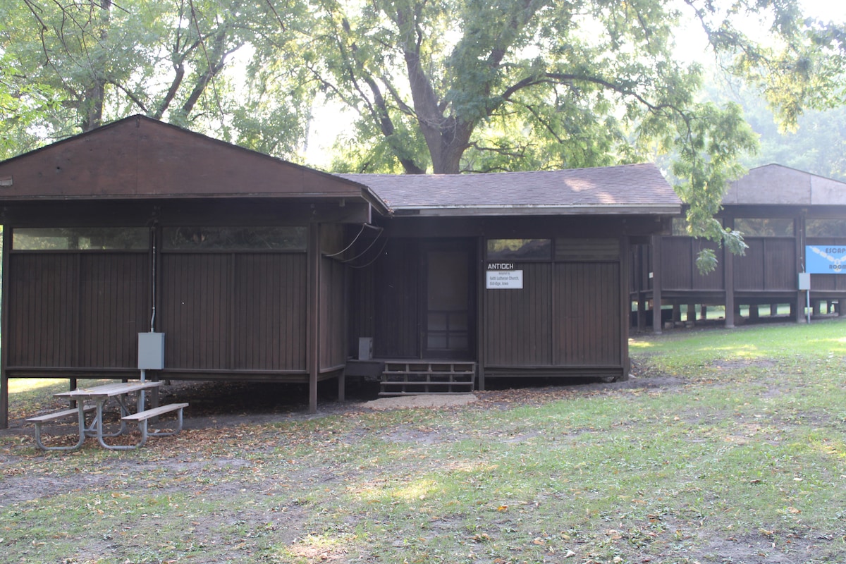 A rustic cabin with dark wooden paneling is situated among trees, featuring a covered porch and a set of steps leading to the entrance. A picnic table is positioned on the grass in front, offering a communal space for outdoor gathering.