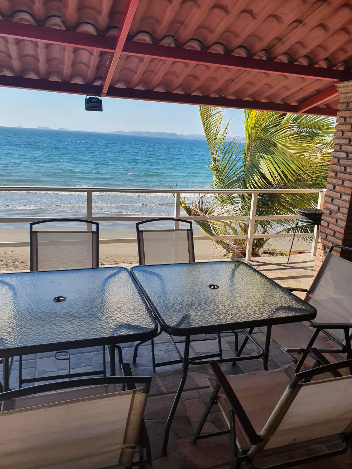 A covered balcony features a glass-top table surrounded by several chairs, offering a view of the ocean and sandy beach below. Palm trees frame the scene, and the sound of gentle waves can be imagined in the background.