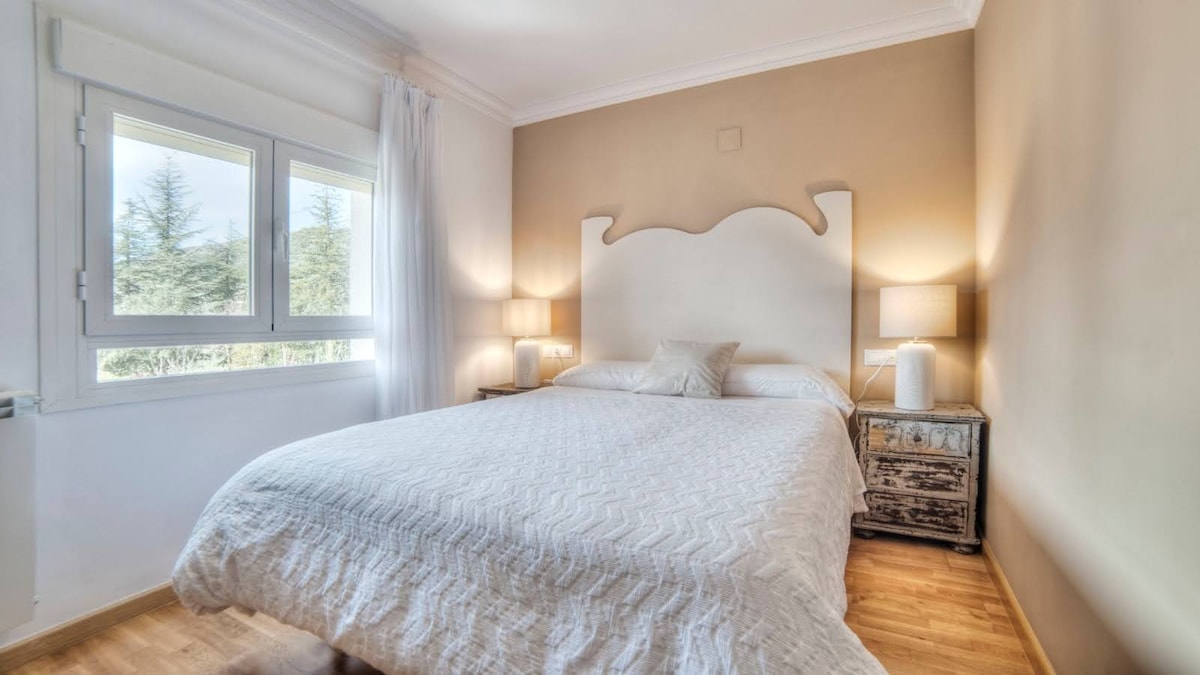 A well-lit bedroom features a spacious bed with a textured white duvet and two bedside tables with lamps. Large windows provide natural light and views of the surrounding greenery, enhancing the calm ambiance of the space.