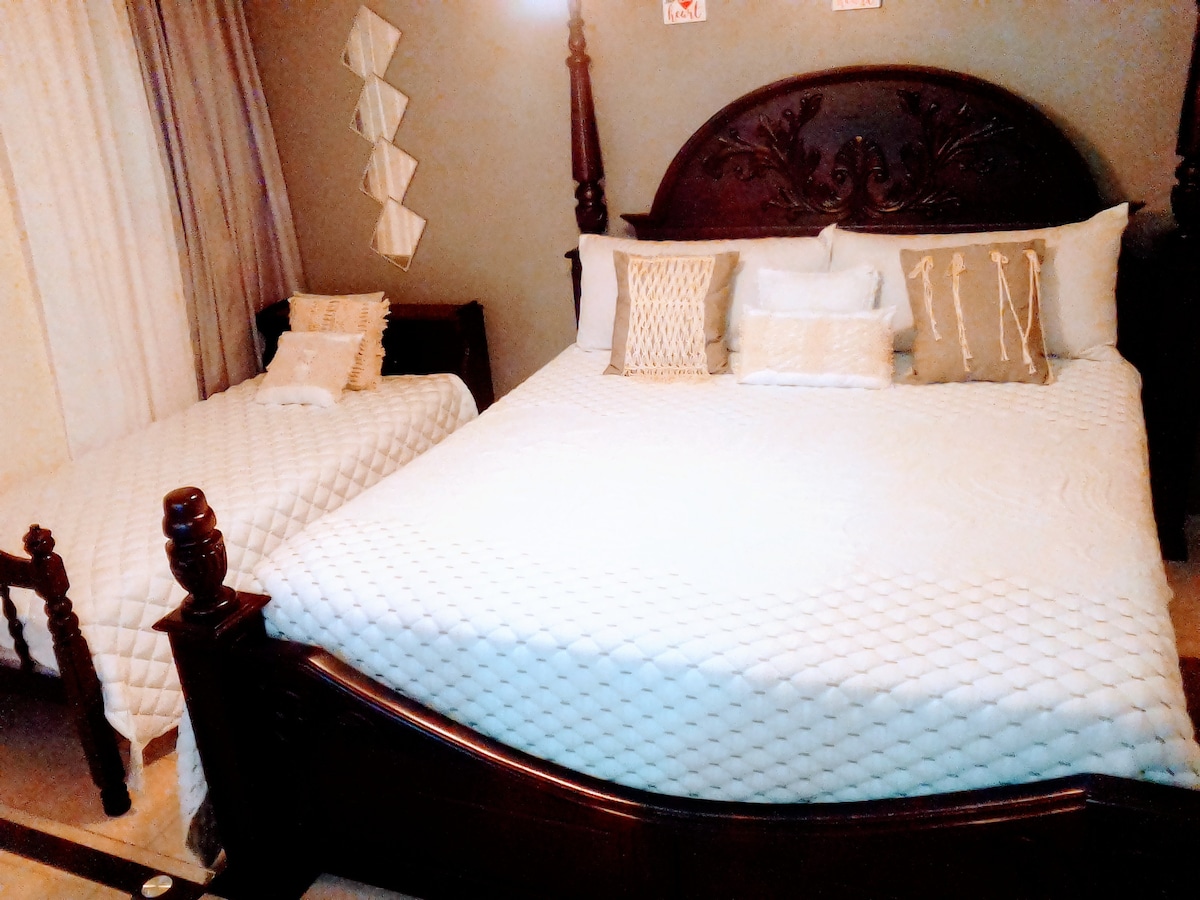 A spacious sleeping area is shown, featuring a large bed adorned with neutral-toned pillows and textured throws. A smaller bed is positioned nearby, offering additional sleeping space. Soft lighting creates a warm ambiance, while light curtains allow some natural illumination.