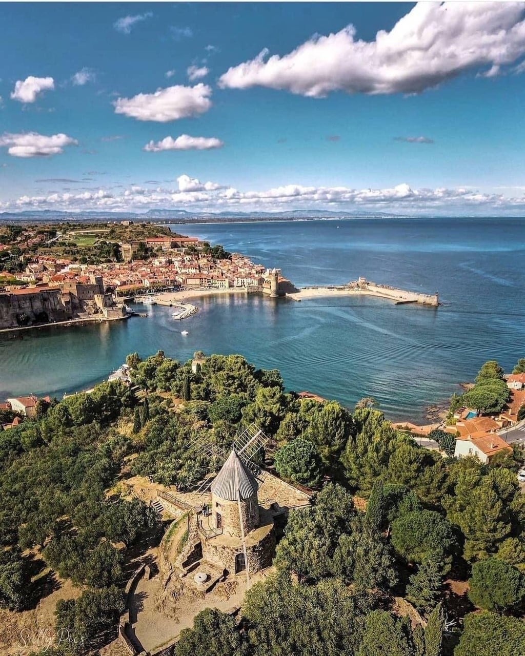 A scenic view captures a coastal landscape featuring a winding harbor with boats. A structure resembling a windmill is positioned among lush greenery, overlooking the water. The horizon is framed by a picturesque town and distant hills under a partly cloudy sky.