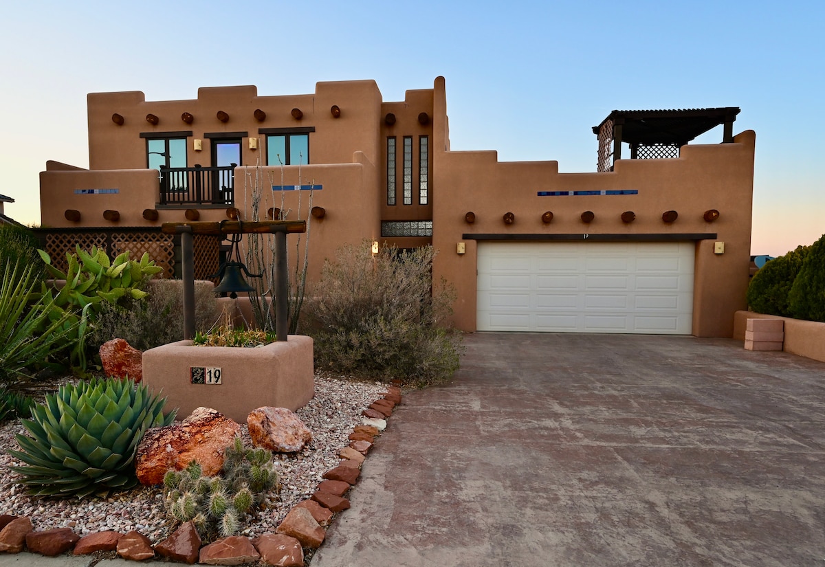 A modern Santa Fe style home features a stucco exterior with wood accents and a spacious two-car garage. The front garden is adorned with desert plants and decorative stones, while a balcony with patio furniture is visible above, offering a view of the surrounding landscape.