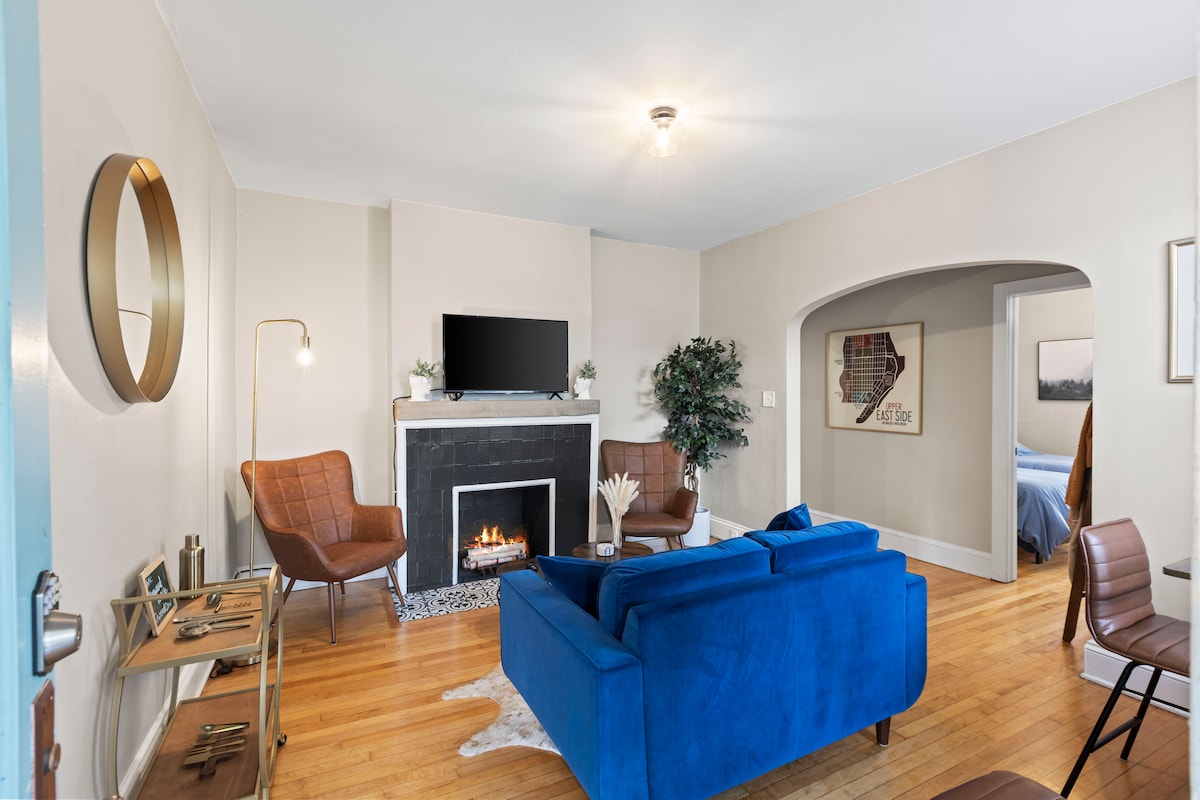 The spacious living area features a blue sofa positioned opposite a decorative fireplace, with a large flat-screen TV mounted above it. Two brown chairs add seating, while a small round table is placed beside them. Natural light enters through a nearby doorway leading to a bedroom.