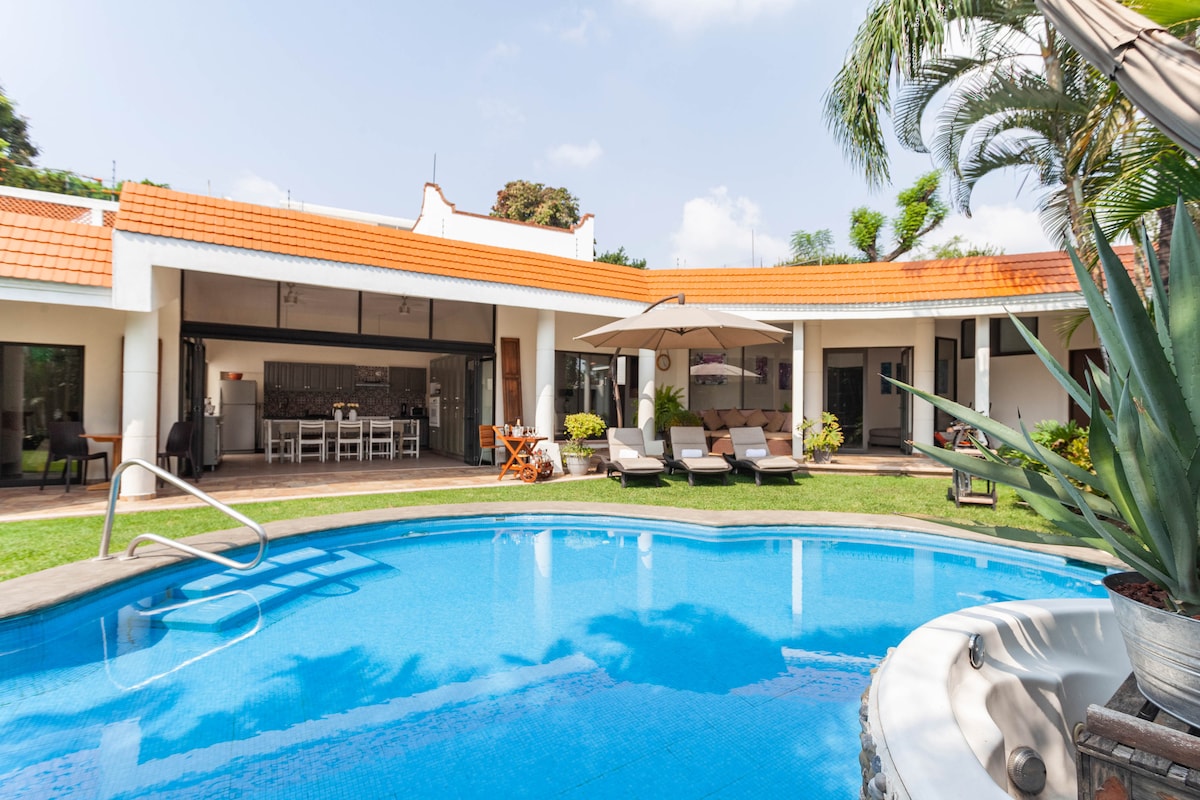 An inviting outdoor area features a circular pool surrounded by lounge chairs and lush greenery. Open-concept living spaces are visible in the background, complemented by modern architecture. Large umbrellas provide shade, while the vibrant blue water reflects the clear sky.
