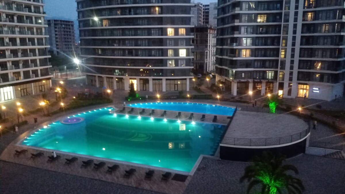 The image captures a serene swimming pool surrounded by modern apartment buildings. Soft blue lights illuminate the water, and lounge chairs are arranged along the edge. Green foliage and decorative lighting enhance the tranquil setting, creating a peaceful ambiance in the evening.