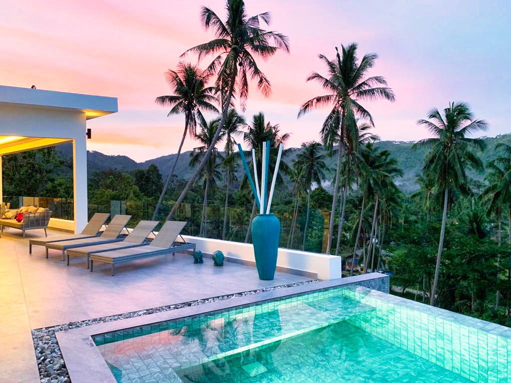 An inviting outdoor area features a serene infinity pool with clear water reflecting the colorful sky. Lounge chairs are arranged alongside the pool, framed by tall palm trees, while a large decorative vase adds a touch of elegance to the space.