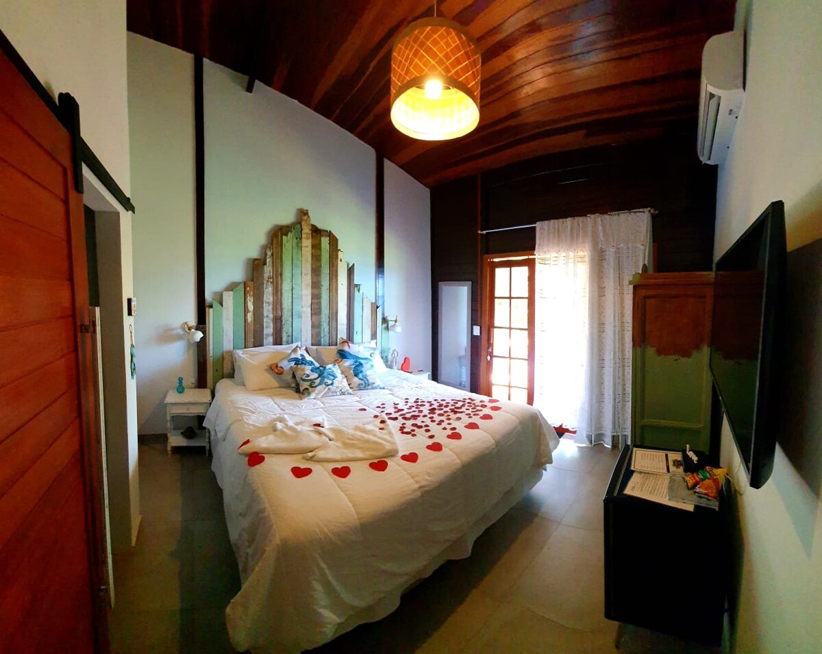 A spacious bedroom features a large bed with a decorative headboard made of wood. Red heart-shaped decorations are arranged on the white bedding. Natural light enters through a door leading to a small balcony, and a ceiling light illuminates the warm wooden ceiling.
