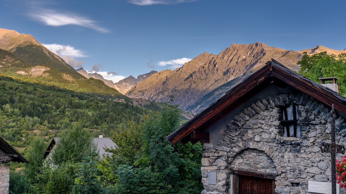 A charming stone structure is nestled among greenery, with a wooden door inviting entry. The backdrop features majestic mountains under a clear blue sky, showcasing the natural beauty of the surrounding landscape.