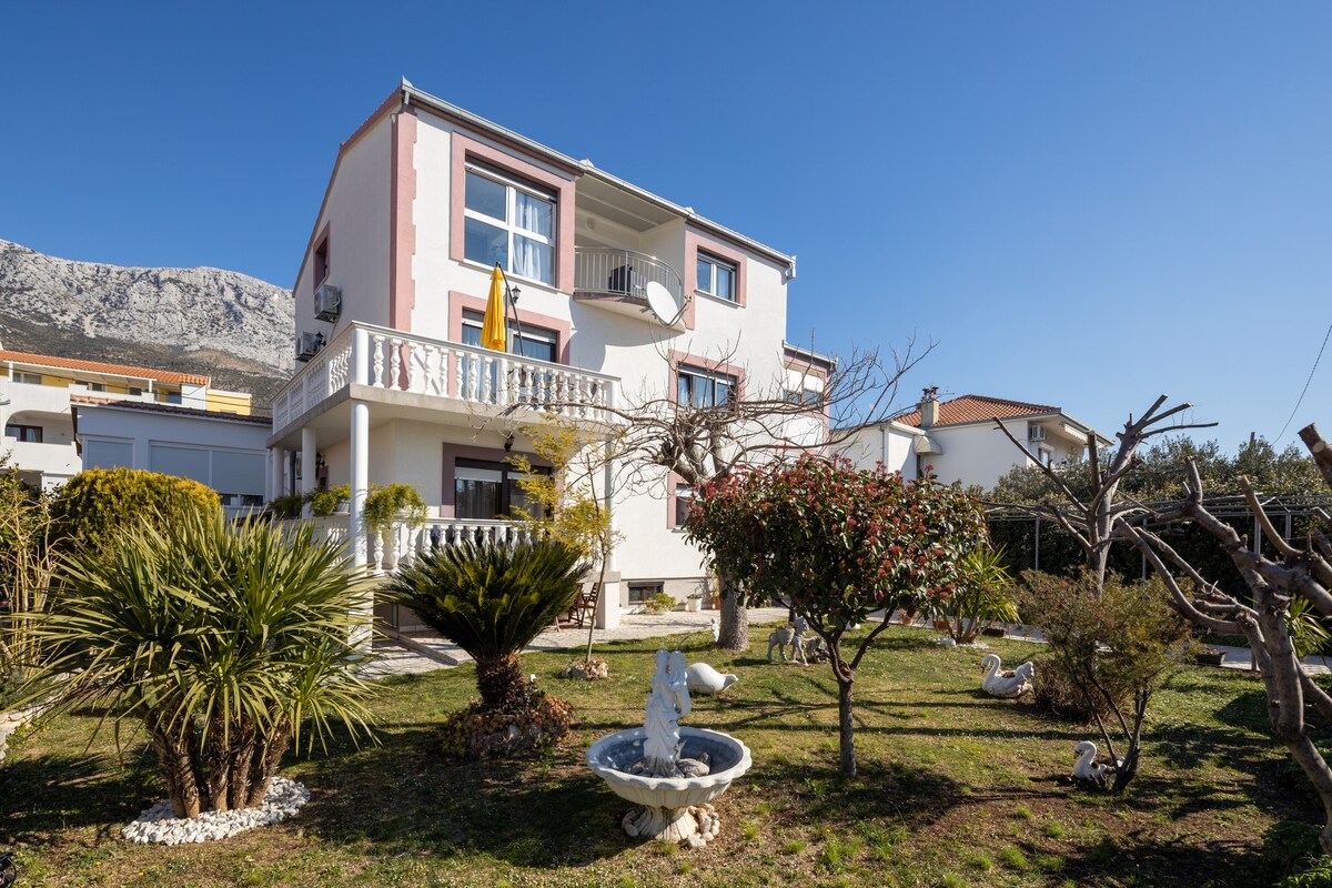 A charming pink building is framed by a well-maintained garden featuring a small fountain and various shrubs. The surrounding area includes palm trees and a gravel pathway that leads to the entrance. A mountain backdrop provides a serene ambience.