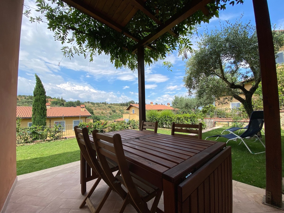 An outdoor dining area is sheltered under a wooden pergola, featuring a large table surrounded by wooden chairs. Lush greenery and trees provide a natural backdrop, while a gently sloping landscape is visible in the distance beneath a partially cloudy sky.