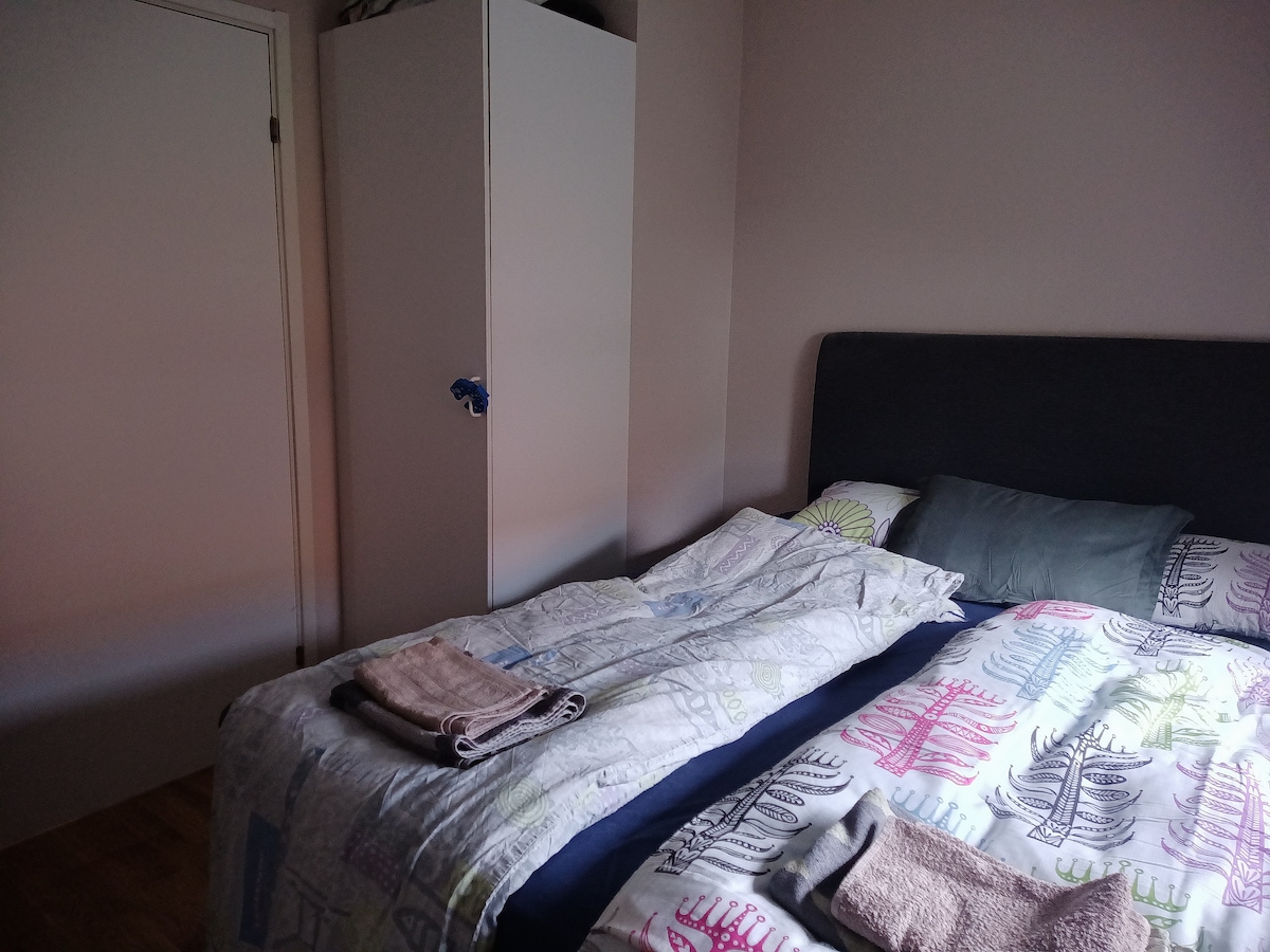 A cozy bedroom is showcased, featuring a spacious bed with patterned bedding and neatly arranged towels. A simple wardrobe is visible in the corner, while soft lighting creates a serene ambiance. The room's neutral walls enhance the overall calmness.