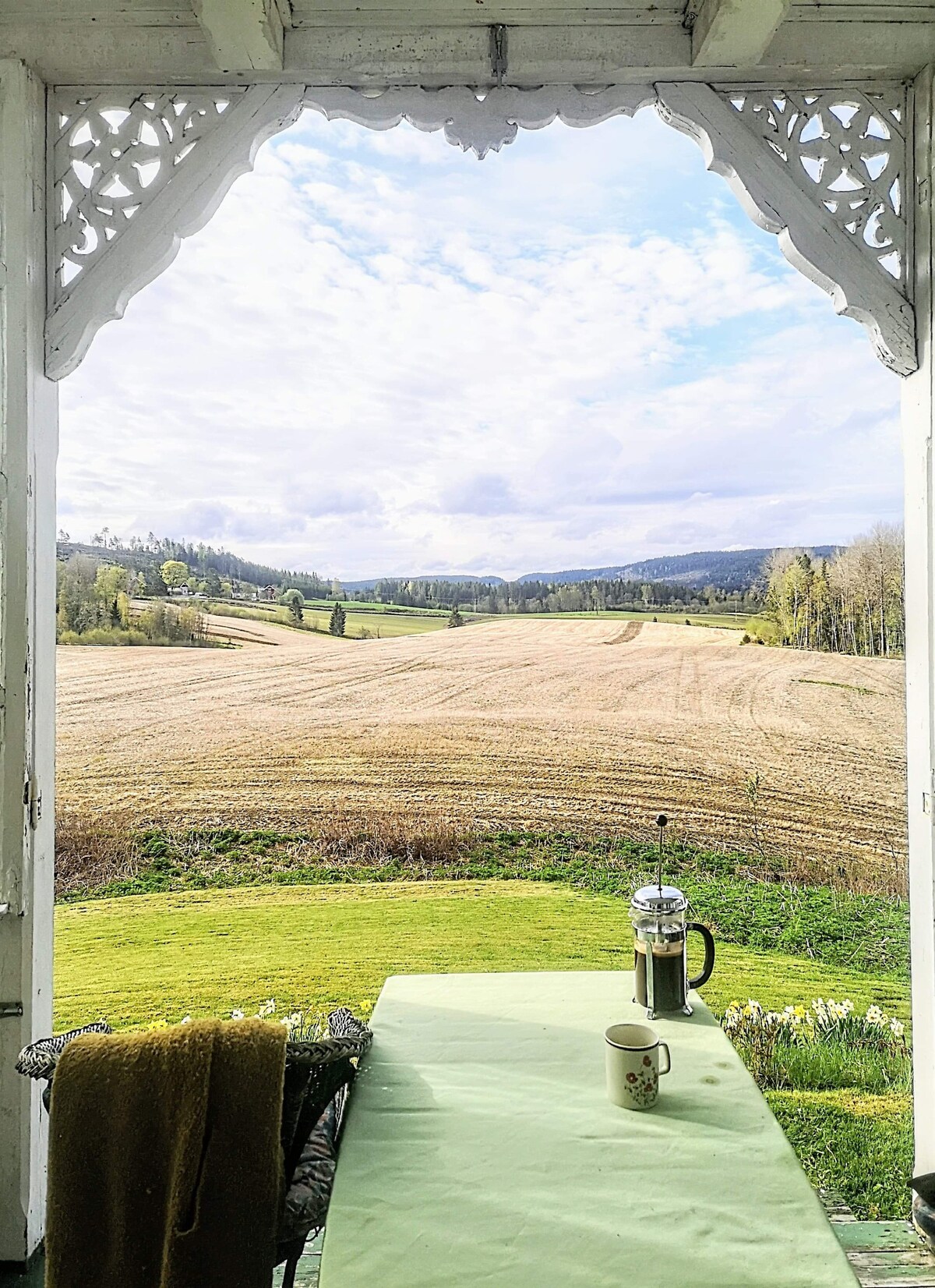 A scenic view is framed by an intricately designed wooden arch, displaying rolling fields and distant hills. A small table with a green tablecloth is set with a coffee press and a mug, creating a serene spot for relaxation.