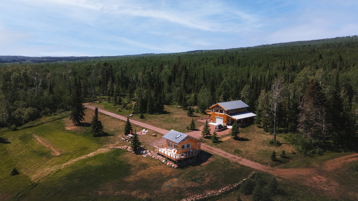Aerial view of the property reveals two modern structures set amidst a vast, green landscape. Surrounding evergreen trees create a natural border, while a gravel driveway meanders through the grounds. Open grassy areas are punctuated with boulders, enhancing the serene, wooded setting.