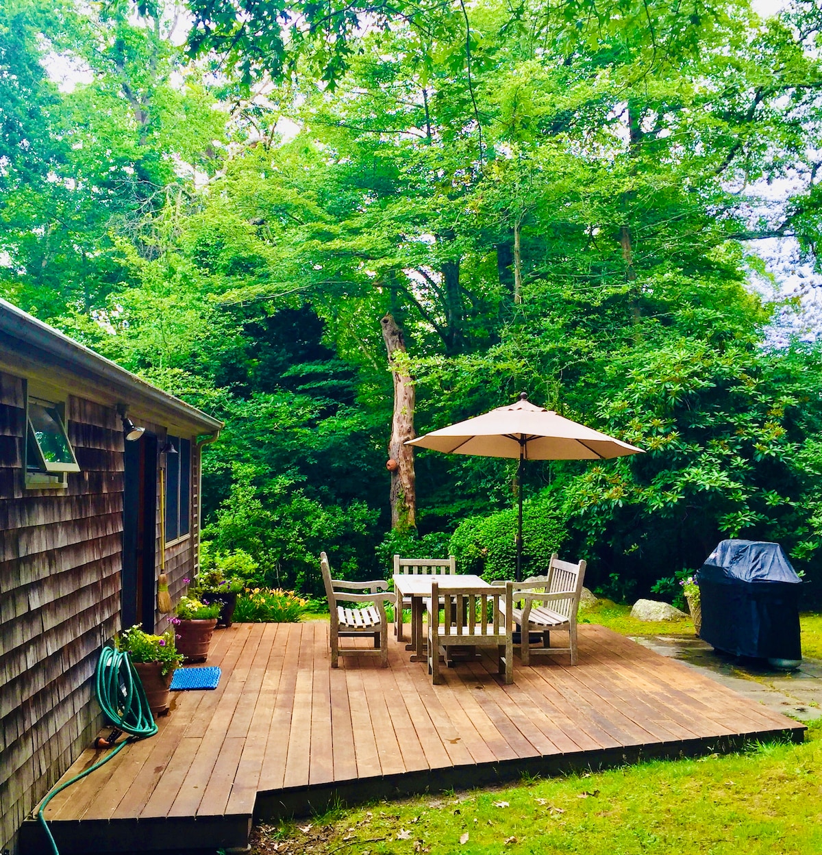 A wooden deck is set amidst lush greenery, featuring a dining table and chairs shaded by a large umbrella. A grill is positioned nearby, providing an ideal space for outdoor meals surrounded by trees.