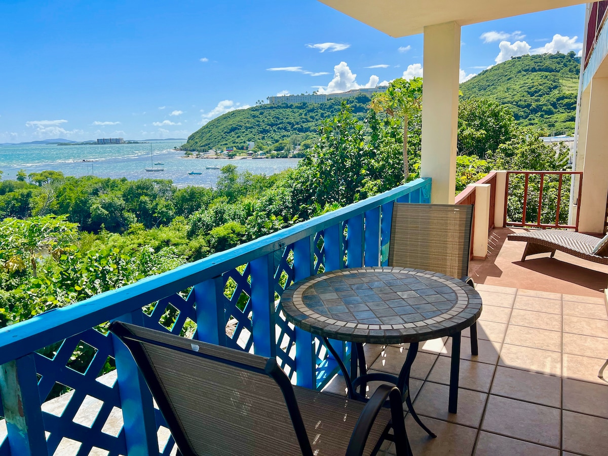 A breezy balcony is displayed with views of the lush green hills and sparkling Caribbean Sea. Two chairs surrounding a round table offer a comfortable outdoor space, complemented by vibrant blue railings and tiled flooring, creating a serene setting for relaxation.