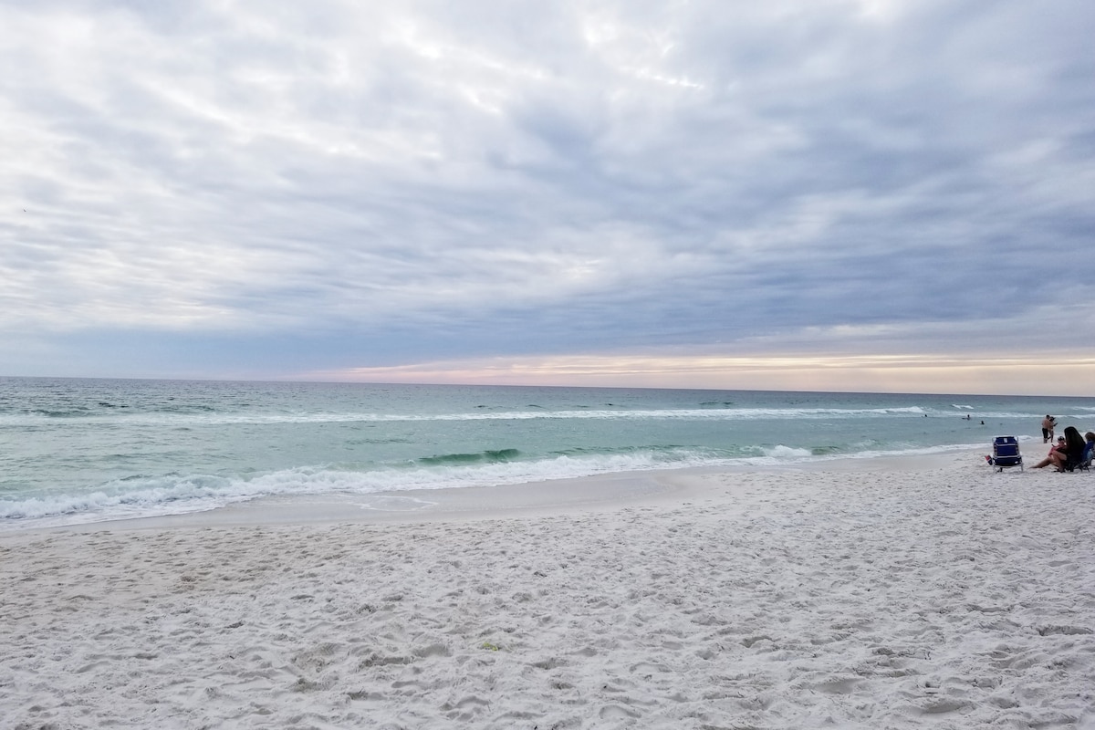 A serene beach scene features soft white sand stretching along the shoreline. Gentle waves are seen lapping against the coast, while the horizon is filled with a soft gradient of pastel colors beneath a partly cloudy sky.