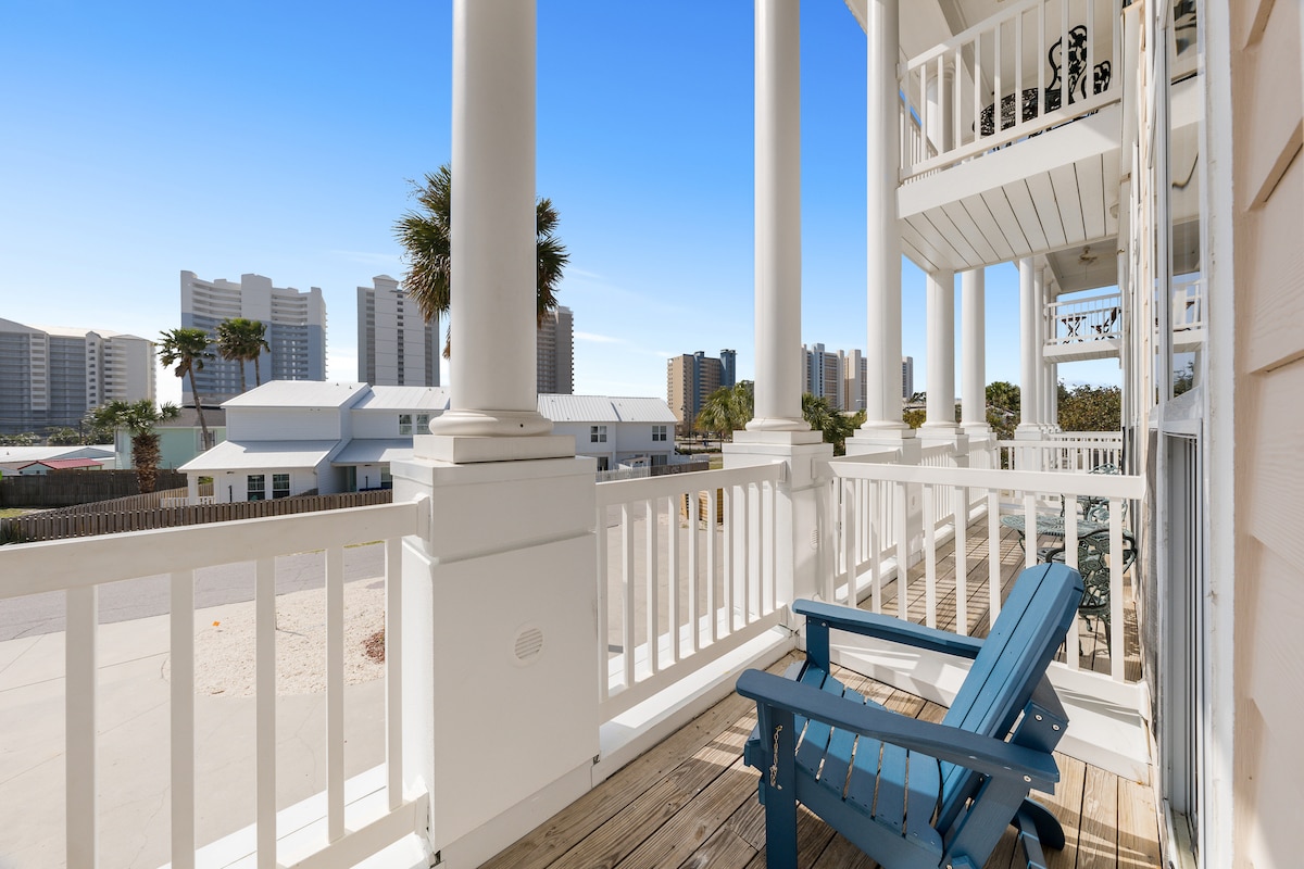 A private balcony is visible, featuring a blue adirondack chair. Tall, white columns support the balcony, with a view of nearby buildings and palm trees in the background. The wooden deck flooring contrasts with the bright blue sky.