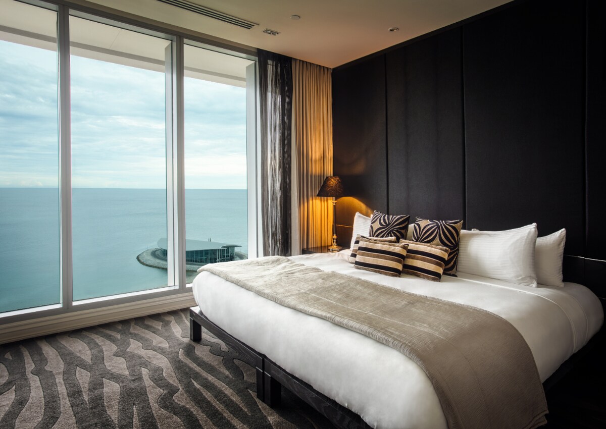 A spacious bedroom features a large bed adorned with textured linens and coordinated pillows. Floor-to-ceiling windows offer expansive views of the sea, while soft lighting creates a serene ambiance. The room is complemented by a stylish area rug with a bold pattern.